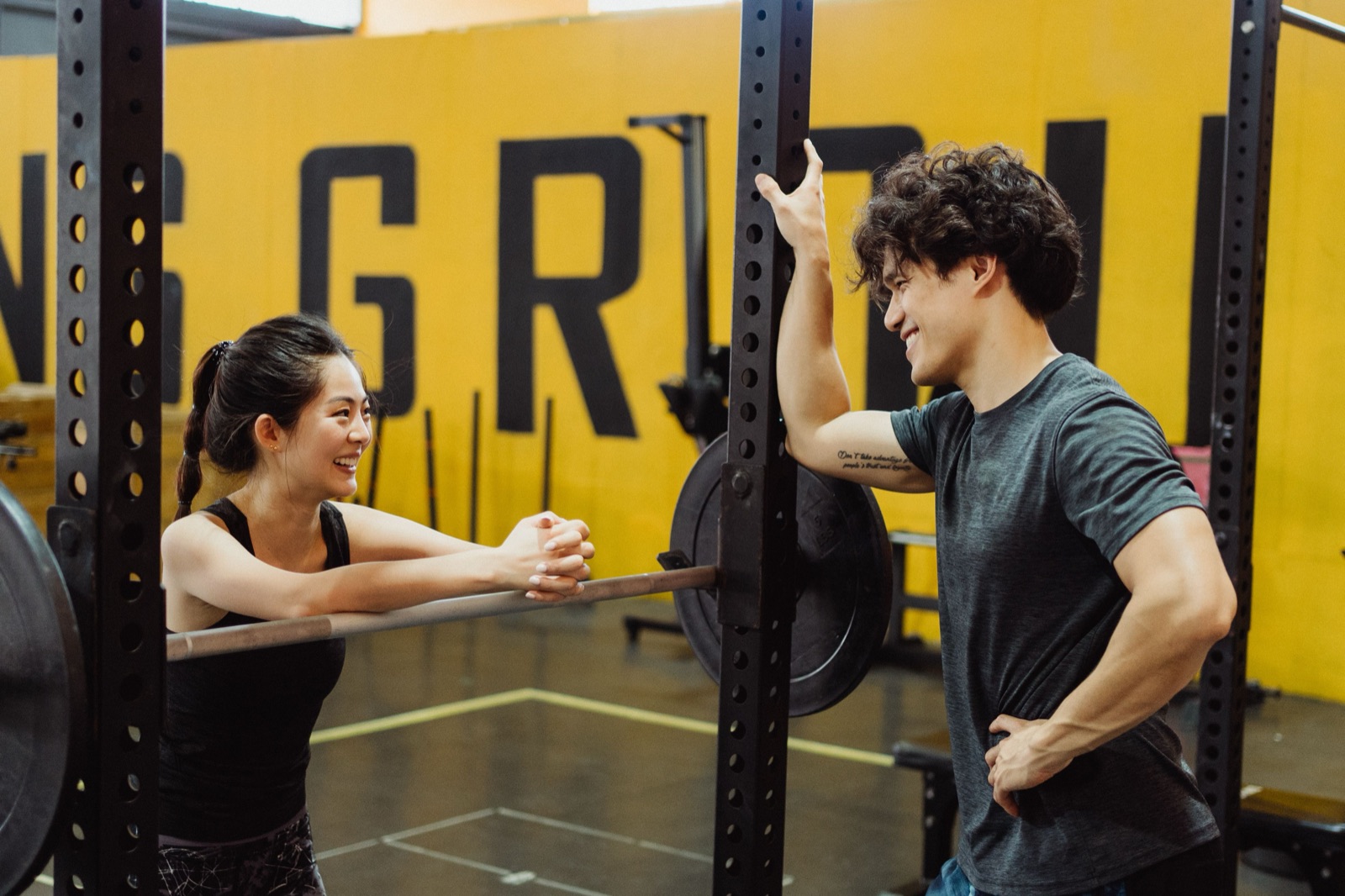 A woman and a man laughing with each other across a squat rack in a gym.