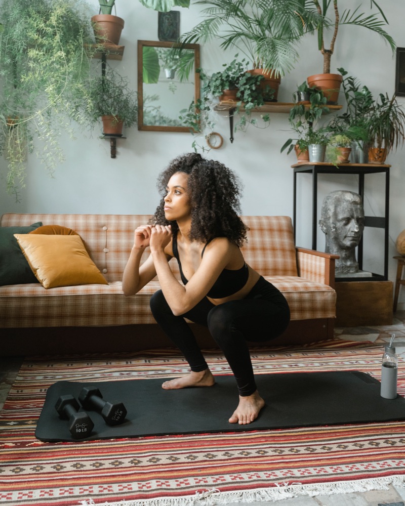 A woman doing a bodyweight squat in a plant-filled living room, a pair of dumbbells on the mat beside her.