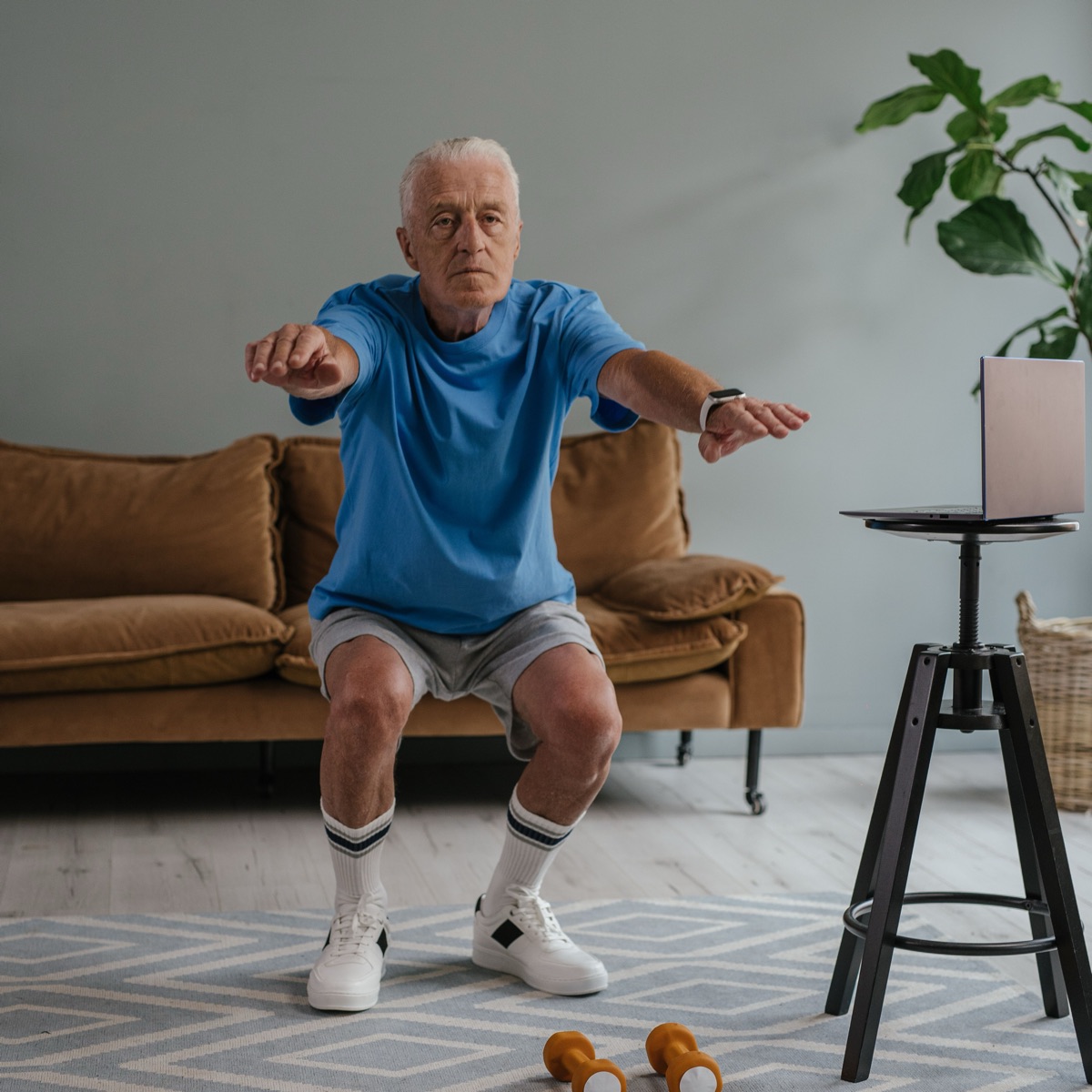 A gray-haired man squatting in his living room, small dumbbells on the floor and a laptop on a stool beside him.